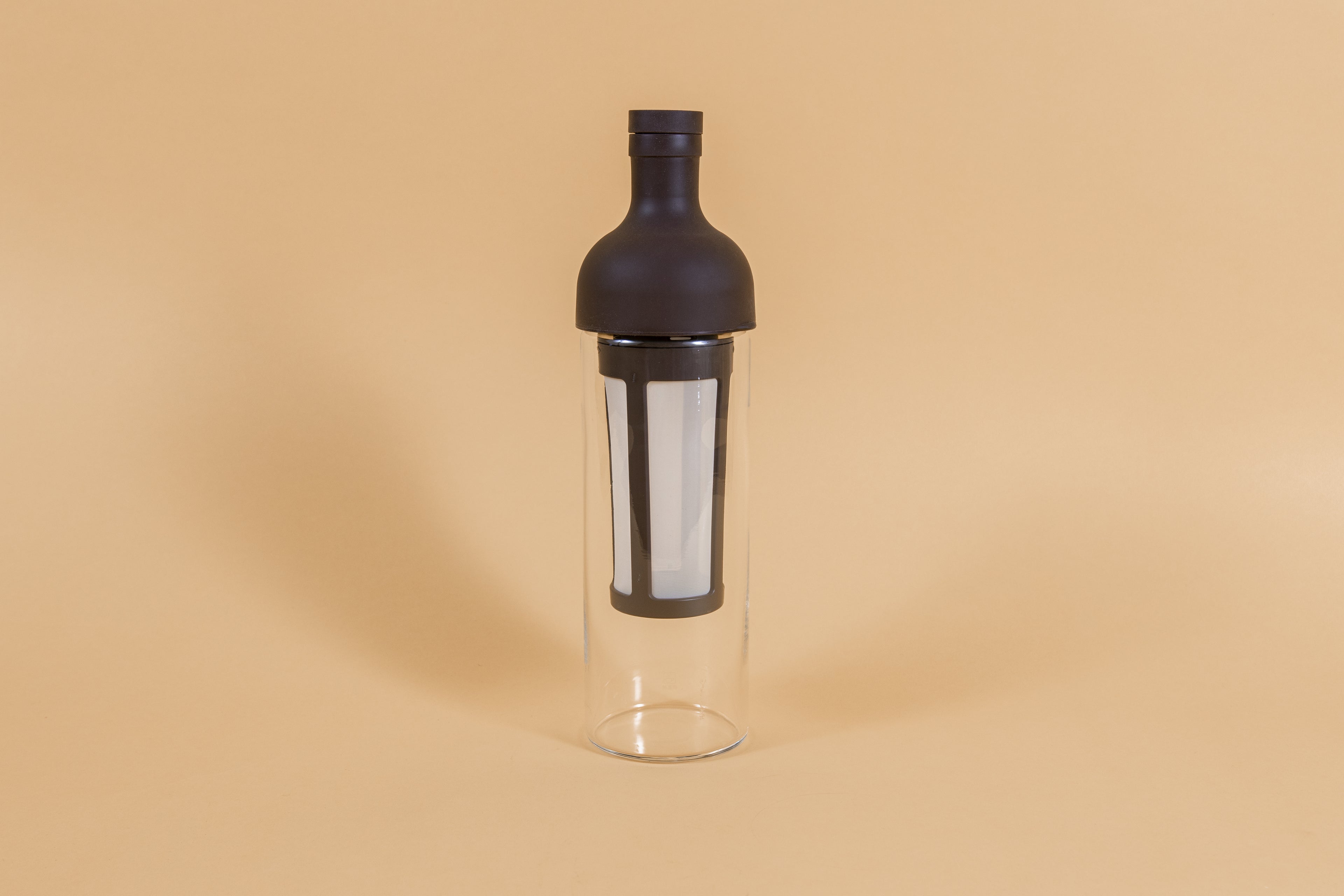 Tall glass container and white nylon mesh coffee filter, with brown rubber wine bottle shaped top on an orange backdrop.