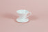 White all plastic cone shaped dripper with handle and round base on a pink backdrop.