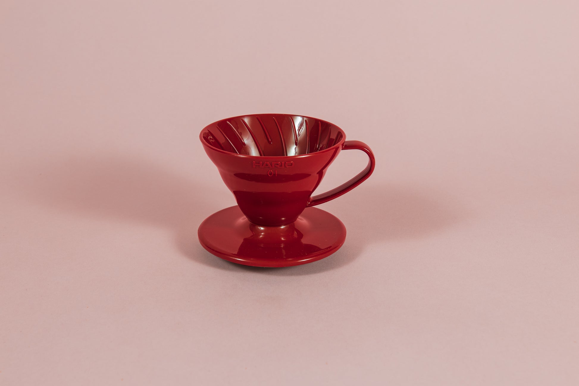 Red all plastic cone shaped dripper with handle and round base on a pink backdrop.