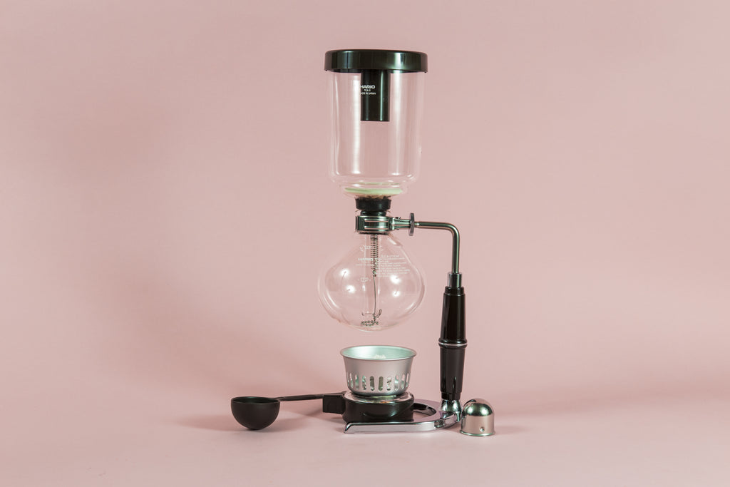 Two piece glass coffee syphon above an alcohol burner attached to a metal and plastic arm with a chrome base with plastic scoop and burner cover on a pink backdrop.