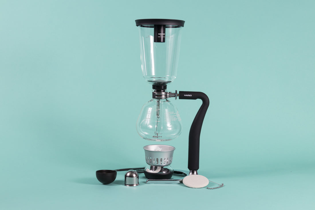 Two piece glass coffee syphon attached to a metal arm with rubber coating and chrome base with alcohol burner, surrounded by plastic scoop, cloth filter and burner lid on a teal backdrop.