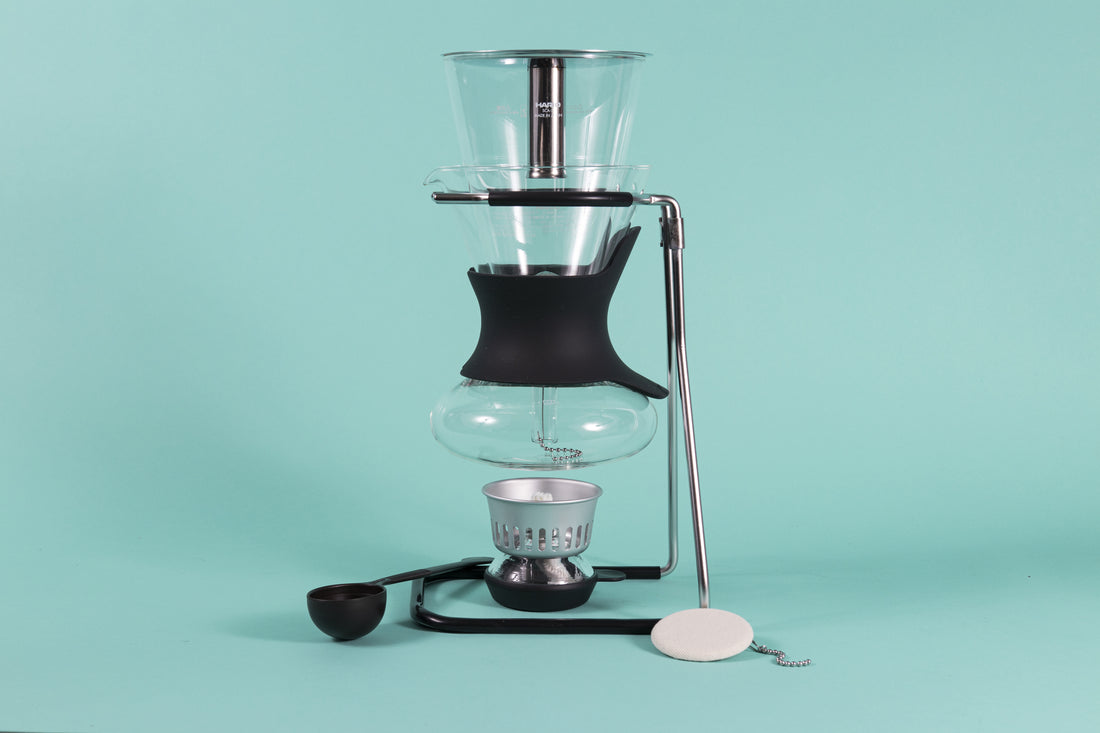 Glass carafe with glass syphon insert resting above an alcohol burner on a chrome stand with rubber sheathes surrounded by plastic scoop and cloth filter on a teal backdrop.