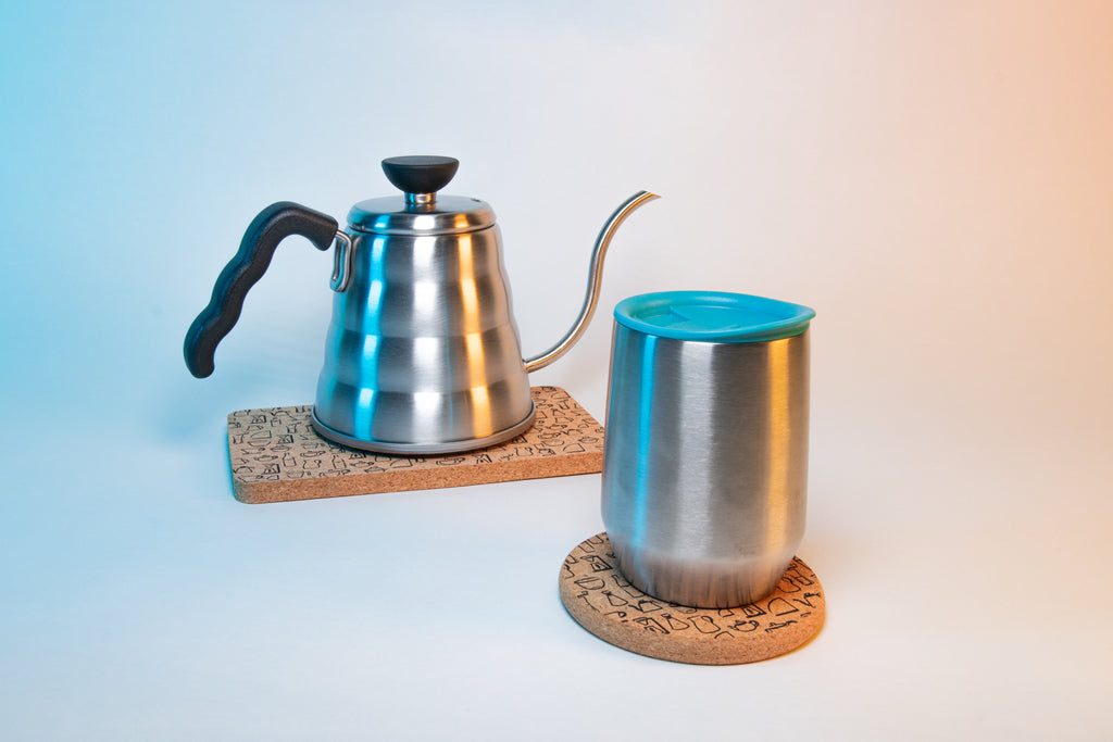 Beehive shaped stainless steal kettle with goose-neck spout, black plastic grip on handle and black plastic lid knob sitting on rectangular cork coaster mat with pattern of Hario products. In front is a stainless steel mug with teal silicone lid sitting on top of a circular cork coaster with pattern of Hario products.