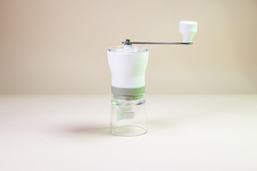 Loose leaf tea grinder with clear plastic base, mesh tea leaf sifter, white powder coated hopper, clear plastic lid, and handle with white powder coated knob.