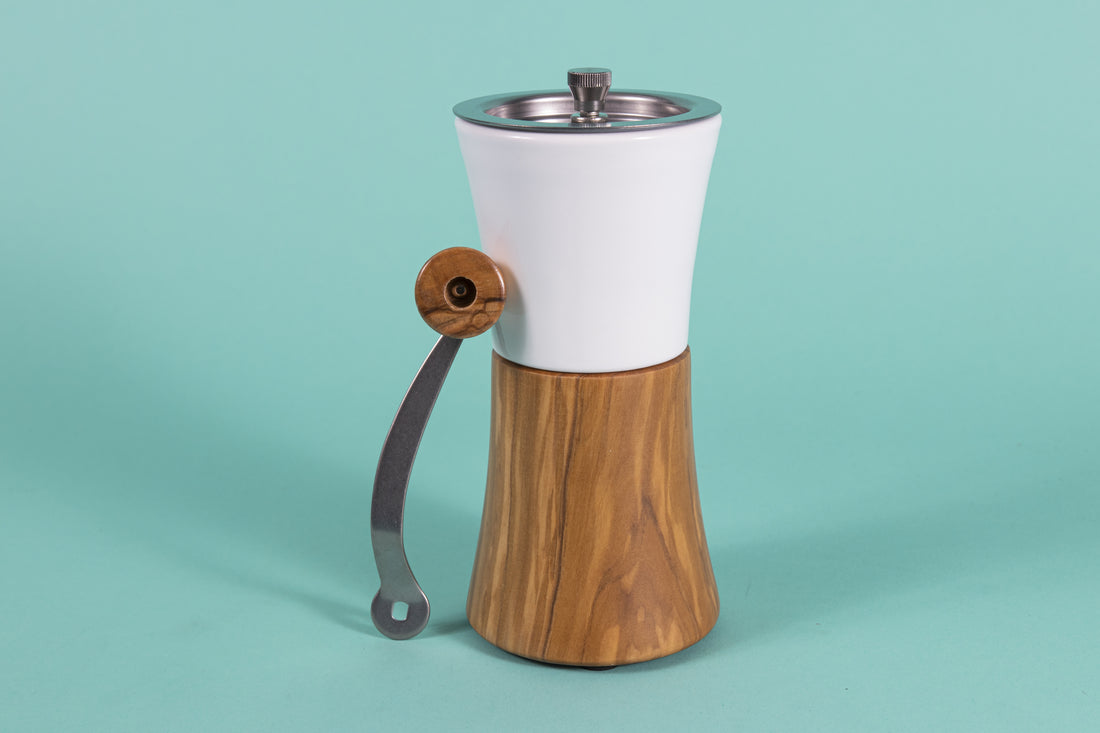 Hour glass shaped coffee hand grinder with olive wood base, white ceramic neck, stainless steel grinder assembly and screw, stainless steel handle with olive wood knob resting against grinder, and set against a blue background.
