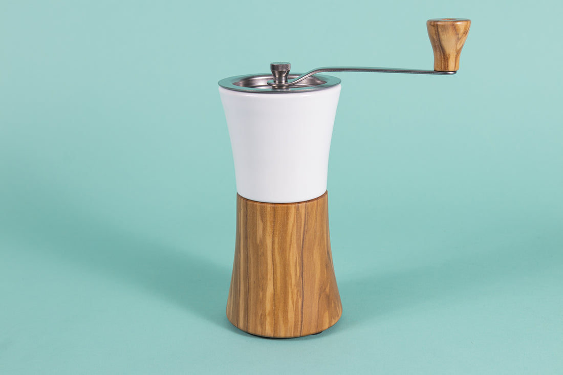 Hour glass shaped coffee hand grinder with olive wood base, white ceramic neck, stainless steel grinder assembly and screw, stainless steel handle with olive wood knob, and set against a blue background.