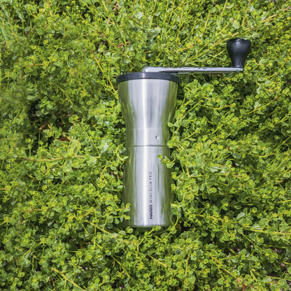Slim silver metal coffee grinder with rubber lid and metal lever with black plastic knob at the end, laying in a bed of green shrubbery.