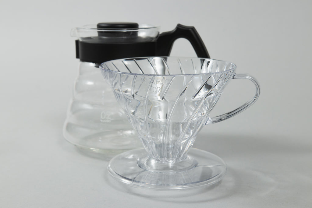 Clear plastic conical dripper with base and handle in front of a glass coffee server with black plastic handle and lid.