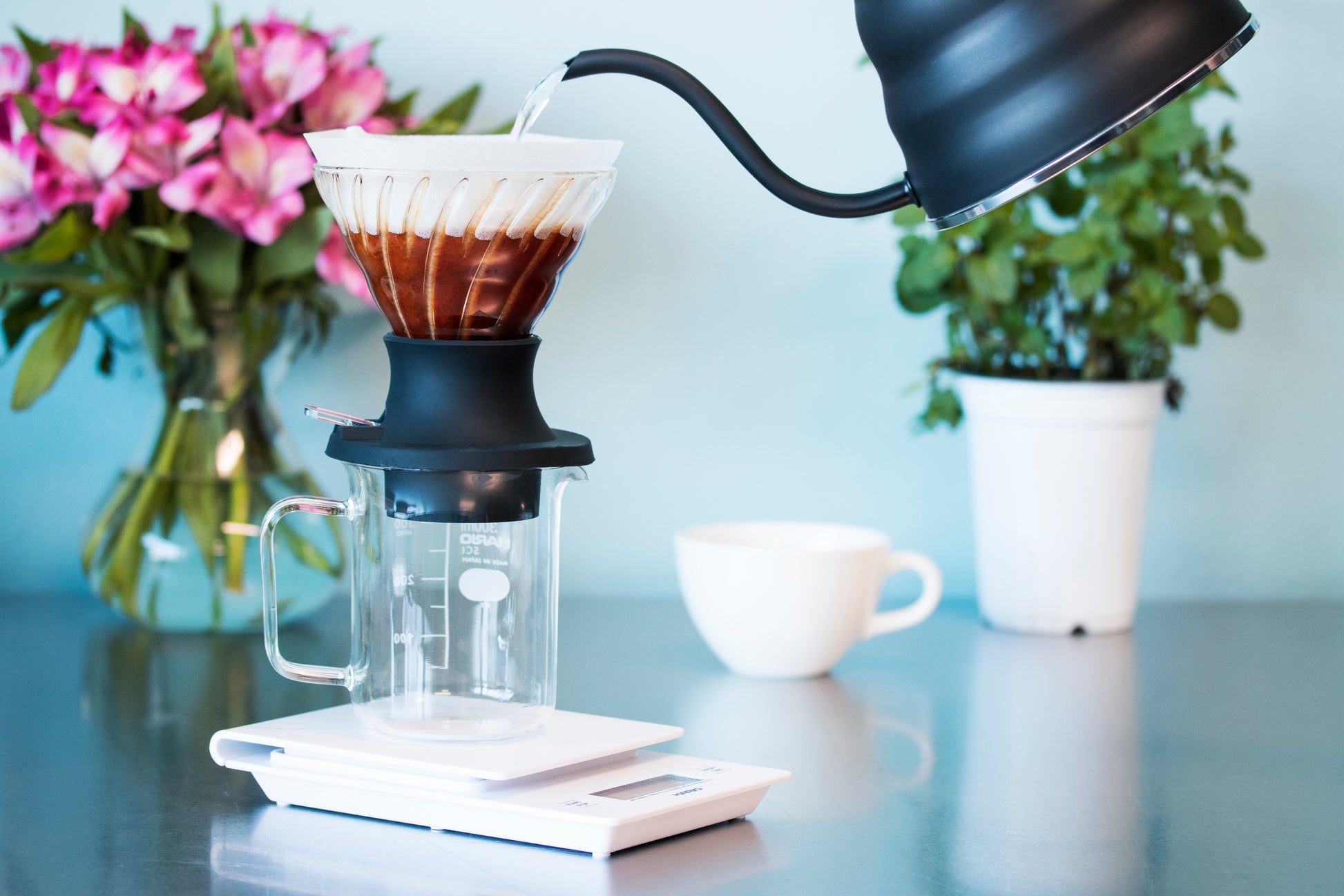 Matte black bee-shaped dripper pouring water into a cone-shaped glass dripper with white paper filter seated in a black silicone base with clear plastic switch and atop a clear glass beaker with white measurement markings and seated on a white plastic scale atop a reflective table top with flowers and a white ceramic coffee cup in the background.