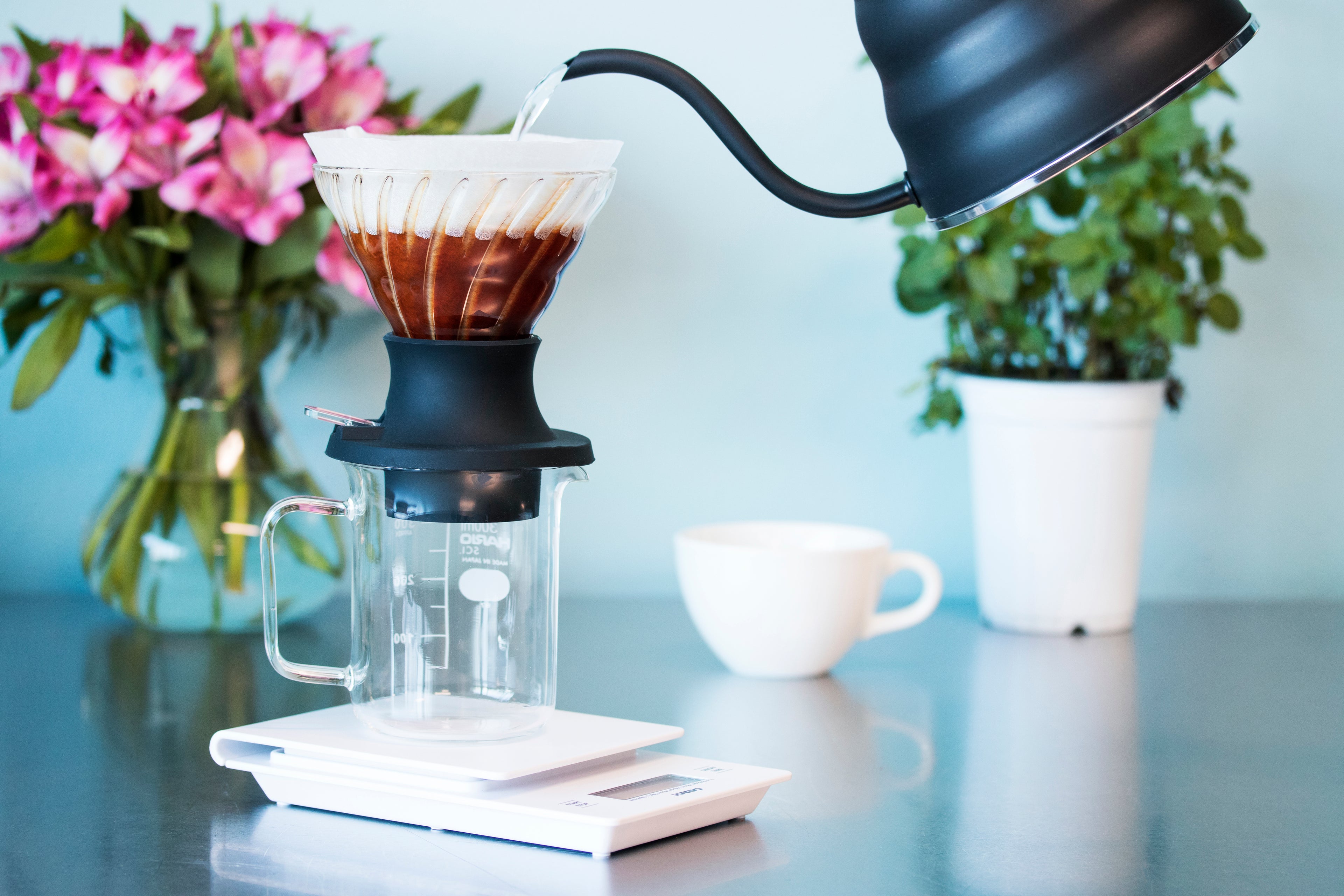 Matte black bee-shaped dripper pouring water into a cone-shaped glass dripper with white paper filter seated in a black silicone base with clear plastic switch and atop a clear glass beaker with white measurement markings and seated on a white plastic scale atop a reflective table top with flowers and a white ceramic coffee cup in the background.