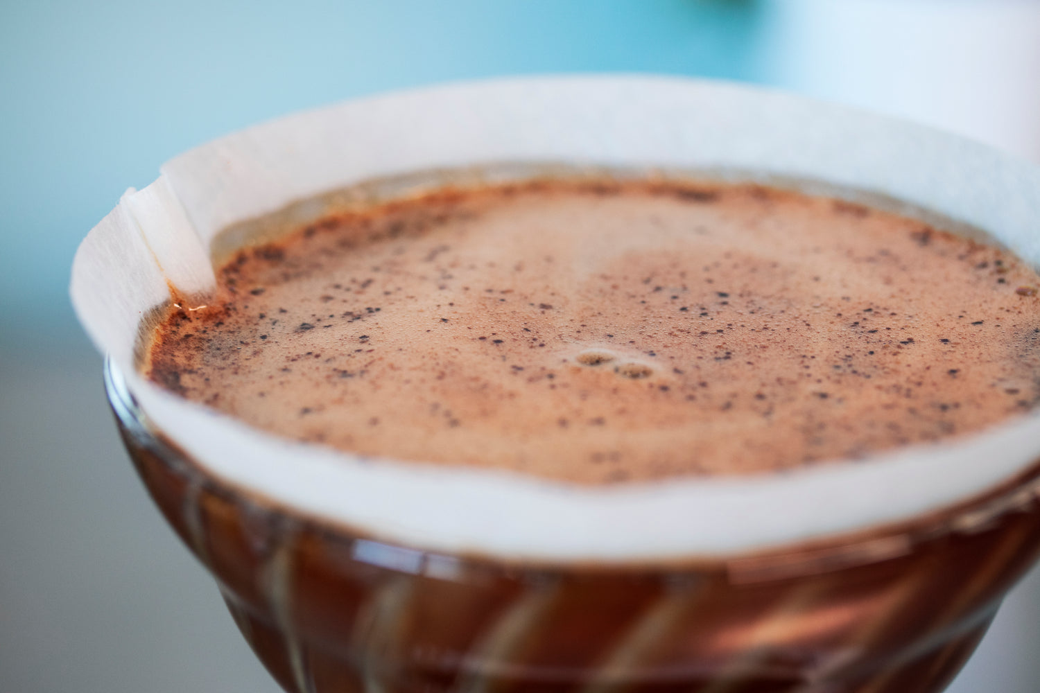 Close up of blooming coffee grinds in a cone-shaped glass dripper with white paper filter.