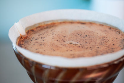 Close up of blooming coffee grinds in a cone-shaped glass dripper with white paper filter.
