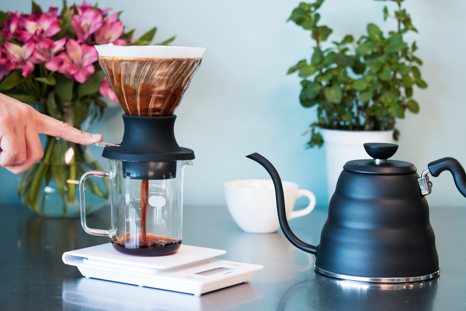 Finger pushing switch to pour brewed coffee from a cone-shaped glass dripper with white paper filter, brewed coffee inside, into a glass beaker with measurement markings in white and seated atop a white plastic scale. Beside the scale is a matte black bee-shaped kettle with flowers and a white ceramic coffee mug in the background.