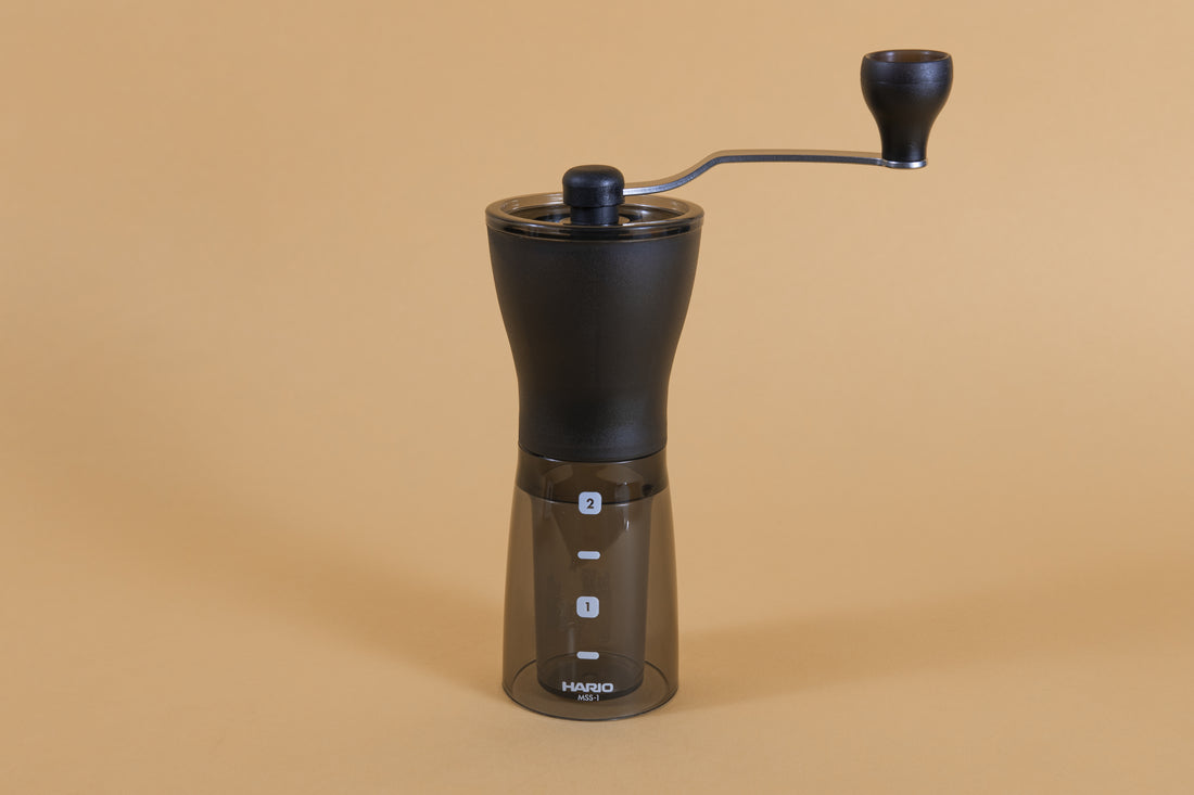 Slim black plastic Coffee Mill with handle atop a dark transparent plastic container with two level markings on an orange backdrop