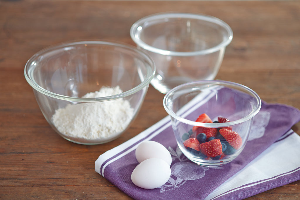 Small, medium, and large glass mixing bowls on a brown wood table and purple and white hand towel. Strawberries and blueberries in the small bowl and flour in the large bowl. Two white eggs on the hand towel beside the small mixing bowl.