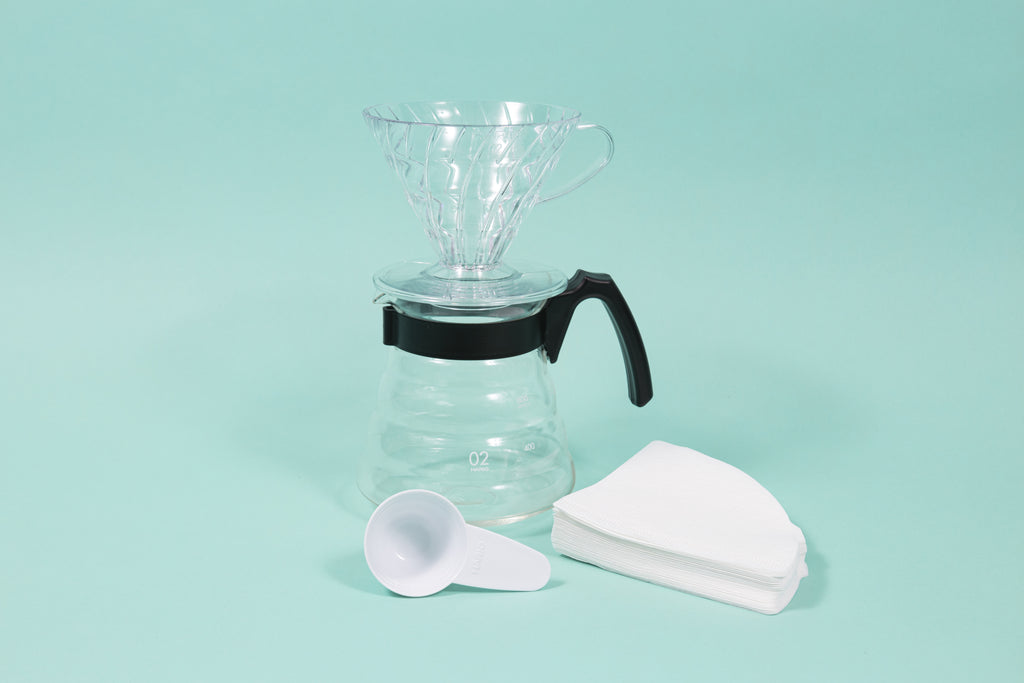 Clear plastic conical coffee dripper atop a glass coffee server i black plastic handle next a stack of filter and white plastic scoop.