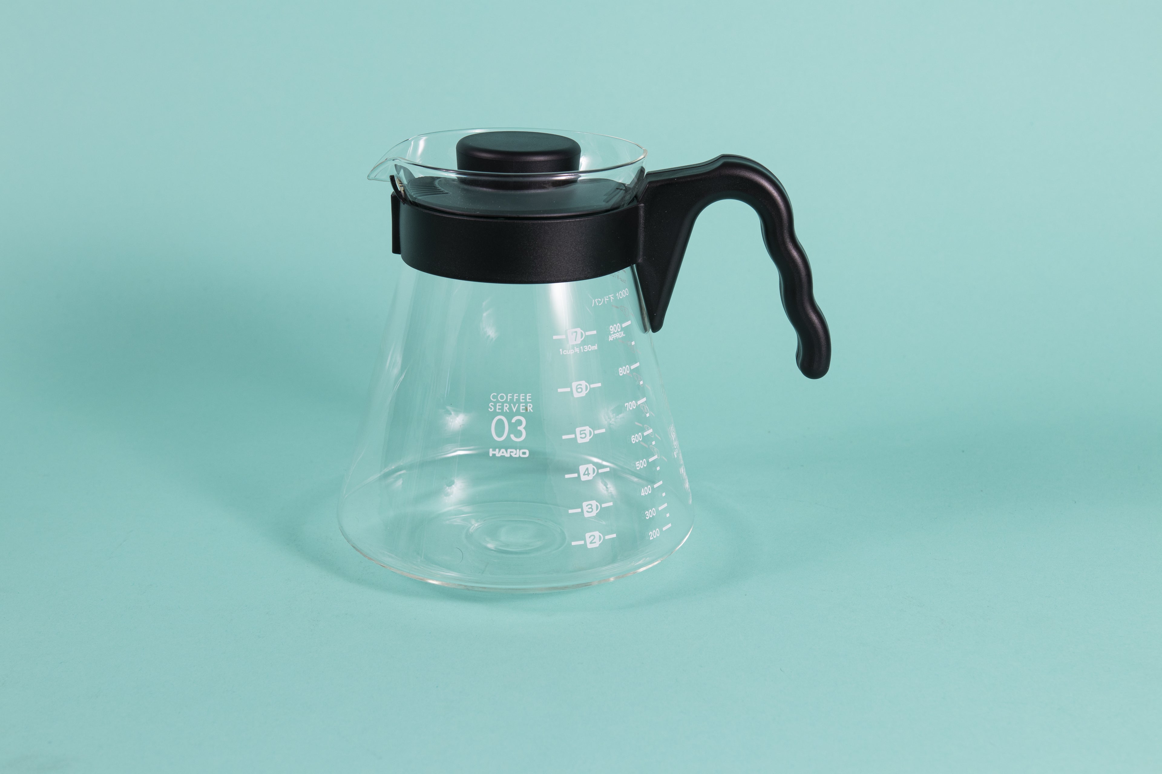 Glass coffee server with white text and level markings with black hard plastic handle and lid on a teal backdrop.