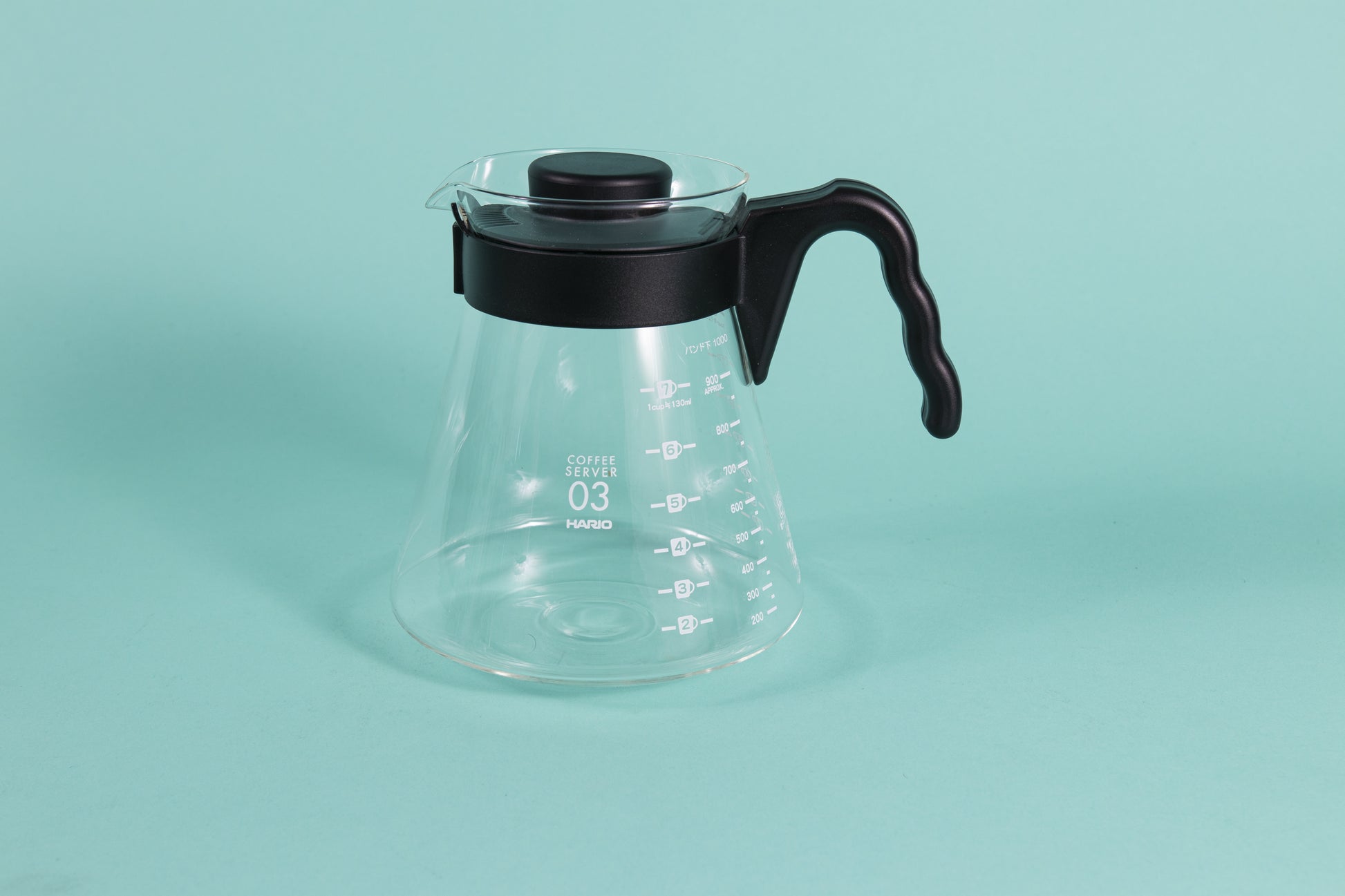 Glass coffee server with white text and level markings with black hard plastic handle and lid on a teal backdrop.