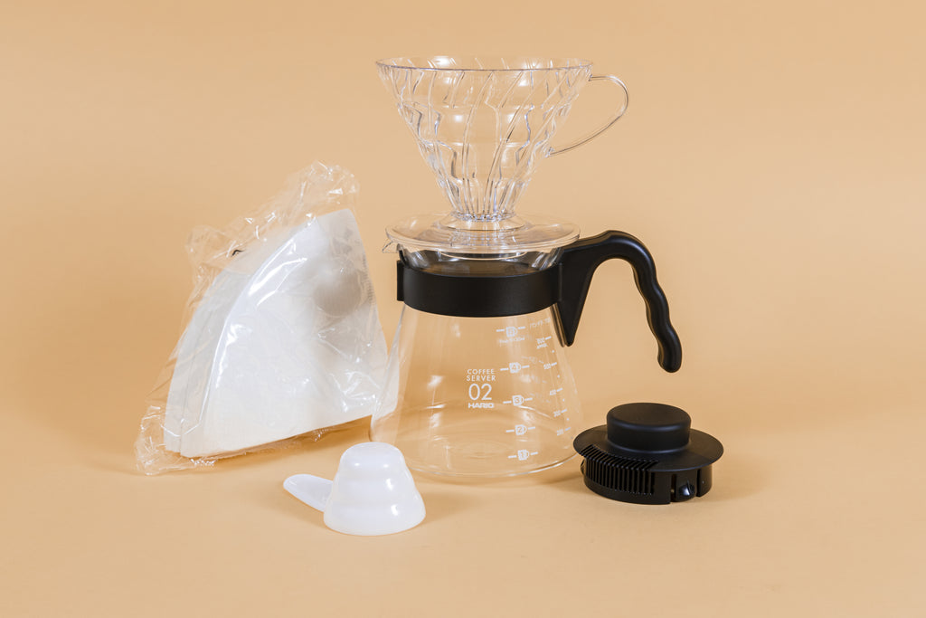 Clear plastic cone shaped dripper sitting on a glass server with black plastic handle next to a pack of white filters and white plastic scoop and black plastic lid.