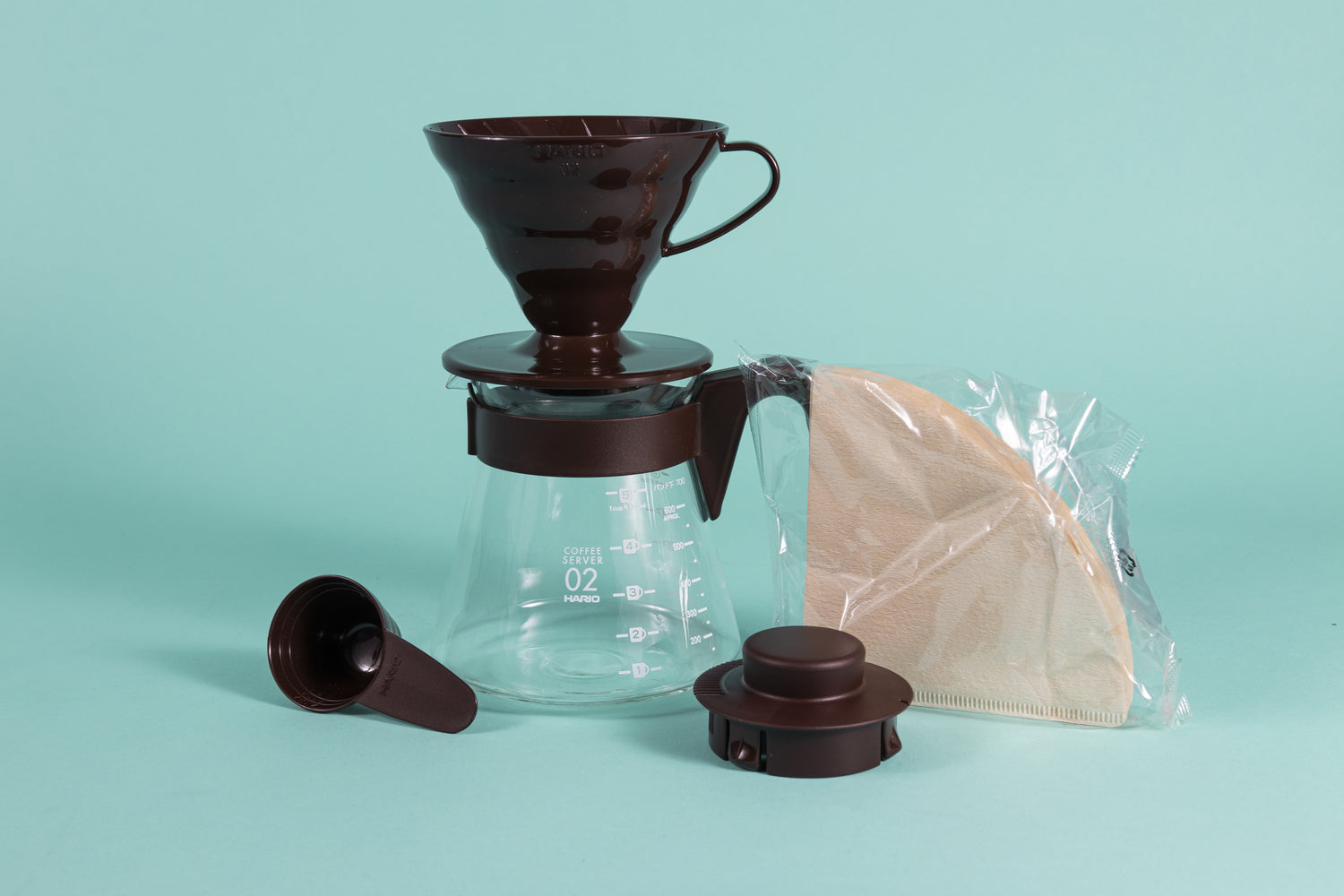 Chocolate brown cone shaped plastic dripper sitting on a glass server with brown plastic handle next to a pack of brown filters and brown plastic lid and scoop.