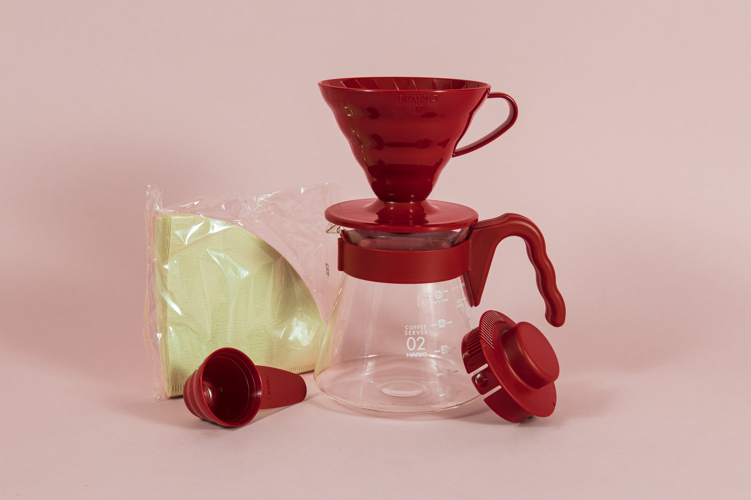 Red all plastic cone shaped dripper sitting on a glass server with red plastic handle next to a pack of brown filters and red plastic scoop and lid.