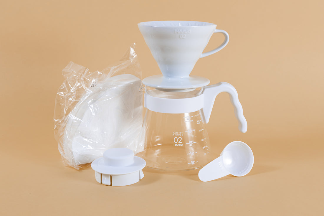 White cone shaped ceramic dripper with handle sitting on a glass server with white plastic handle next to pack of white cone filters white plastic lid and scoop.