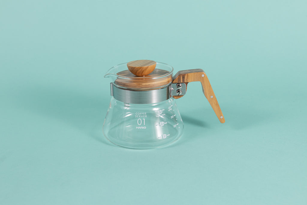 Glass coffee server with white text and markings with a brushed metal collar and wooden handle and lid on a teal backdrop.