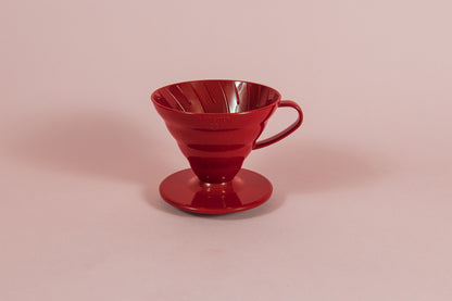 Red all plastic cone shaped dripper with handle and round base on a pink backdrop.