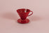 Red all plastic cone shaped dripper with handle and round base on a pink backdrop.