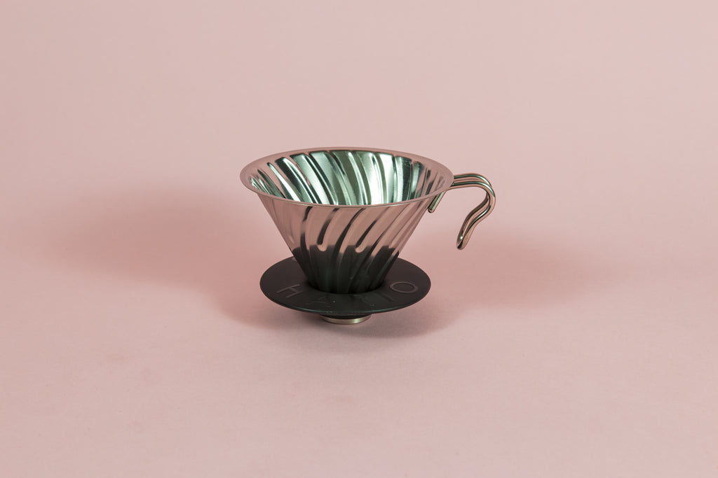 Silver metal cone shaped dripper with ribs and metal handle with a round black rubber base on a pink backdrop.