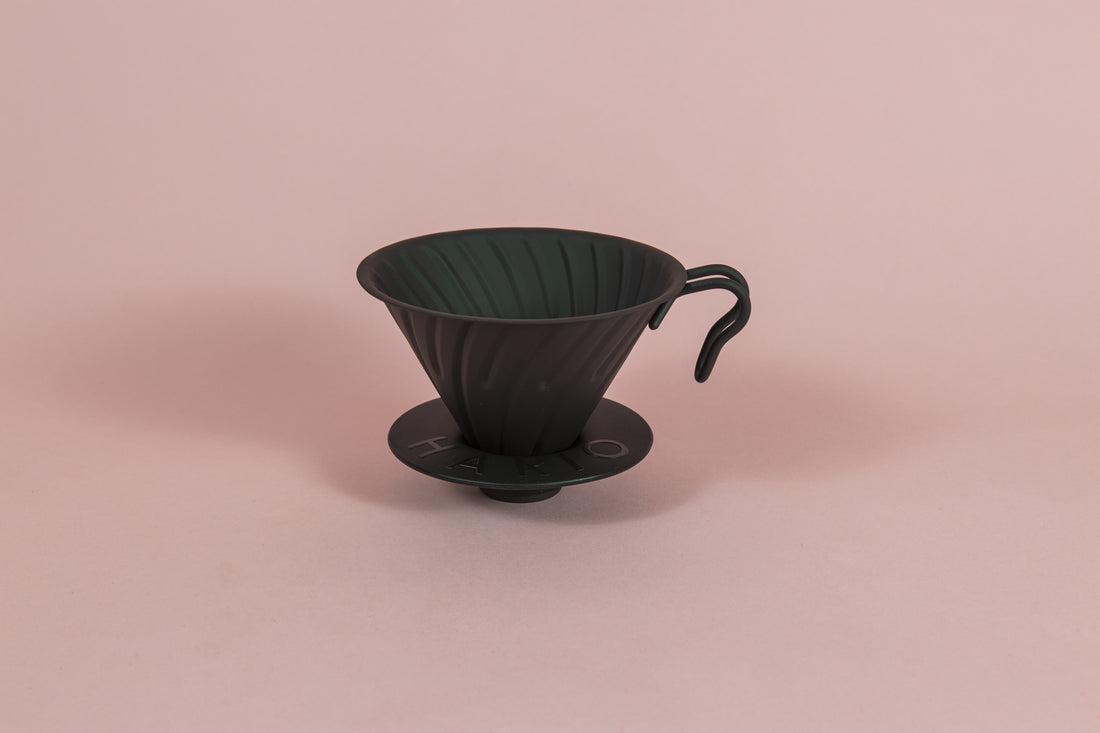 Matte black metal cone shaped dripper with ribs and metal handle with a round black rubber base on a pink backdrop.