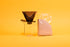 Translucent black plastic cone shaped dripper with bezier ridges and oval shaped black plastic base, set on a glass beaker server with glass handle with a cellphone packaged filters. set on a  yellow background.