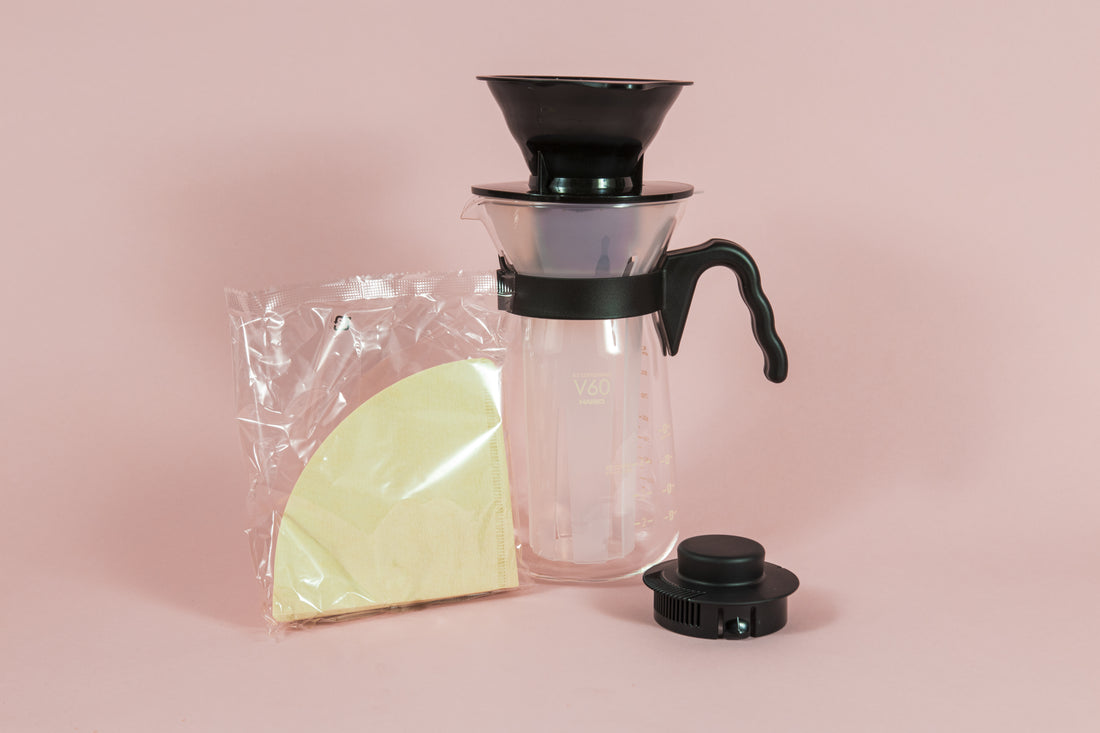 Tall glass coffee decanter with black plastic handle with a frosted plastic insert and black plastic cone dripper on top with black plastic server lid and pack of brown filter.