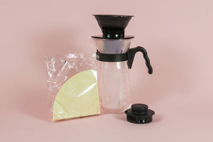 Tall glass coffee decanter with black plastic handle with a frosted plastic insert and black plastic cone dripper on top with black plastic server lid and pack of brown filter.