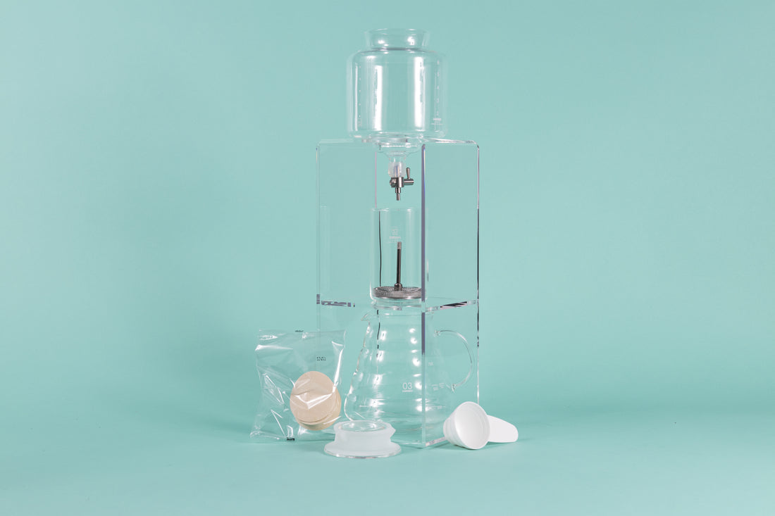 Tall clear acrylic tower stand with water reservoir and drip spout sitting over a glass cylinder with metal mesh filter for coffee grounds, with all glass coffee server with handle underneath and paper filter pack, server lid, and white plastic scoop on a teal background