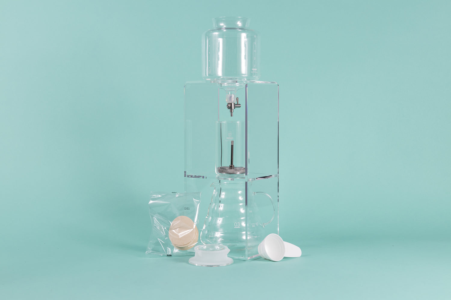 Tall clear acrylic tower stand with water reservoir and drip spout sitting over a glass cylinder with metal mesh filter for coffee grounds, with all glass coffee server with handle underneath and paper filter pack, server lid, and white plastic scoop on a teal background