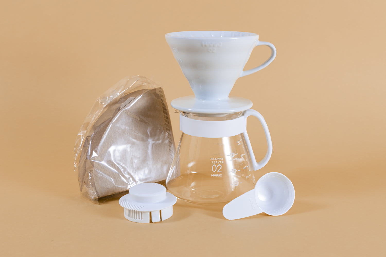 White cone shaped ceramic dripper with handle sitting on a glass server with white plastic handle next to pack of brown cone filters white plastic lid and scoop.