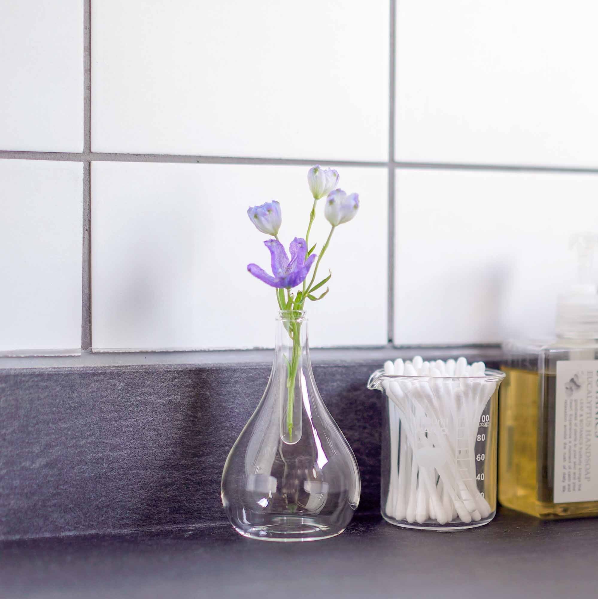 Tear drop shaped glass diffuser with purple and white flowers seated on a grey bathroom countertop with white tile behind and beside a glass beaker with cotton swabs and yellow hand soap.