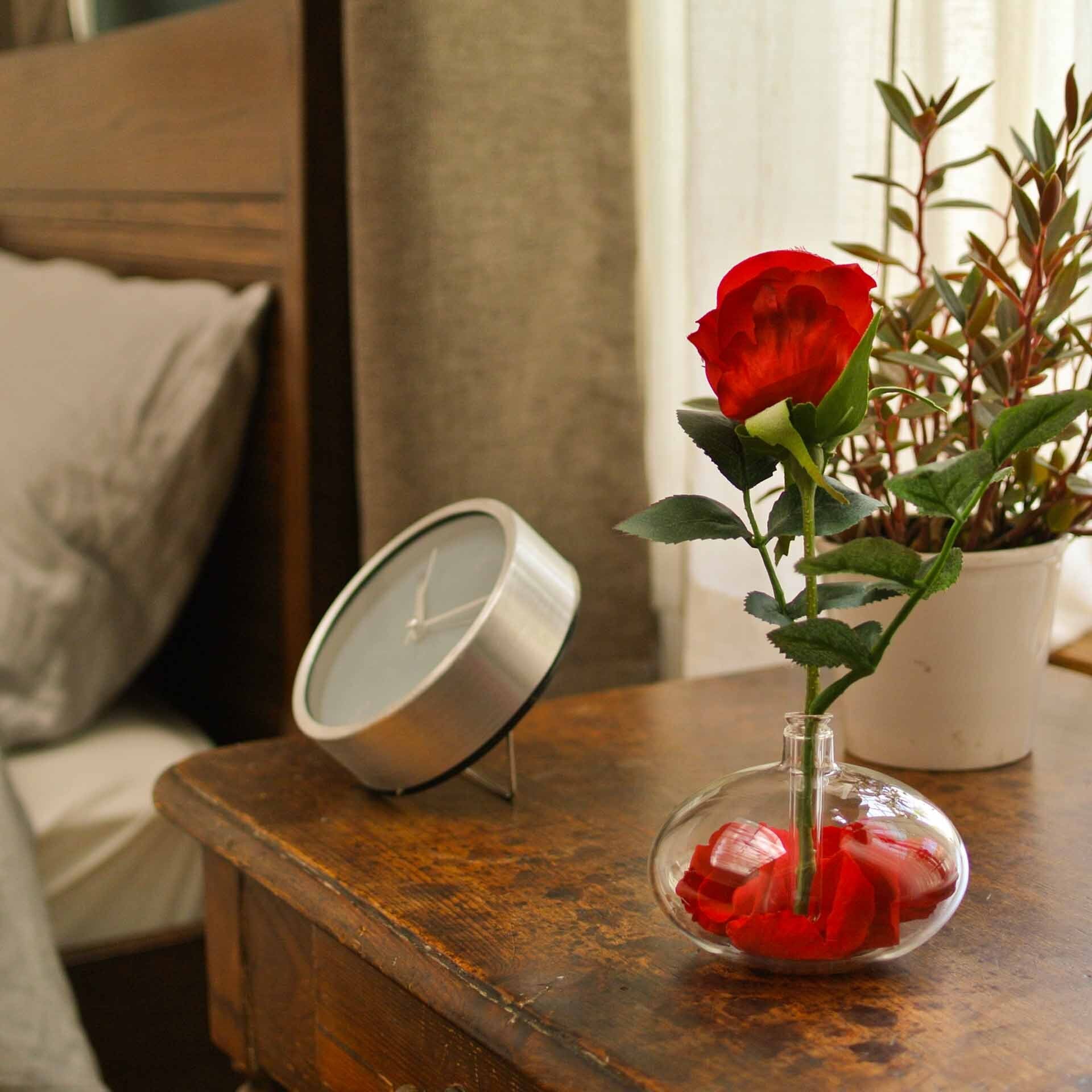 Red rose with red rose petals inside an oval shaped glass diffuser with natural wood wand and round knob, seated on a mahogany nightstand with silver analog clock and white potted plant with a bed with grey comforter, pillow, and white linen sheet in the background..