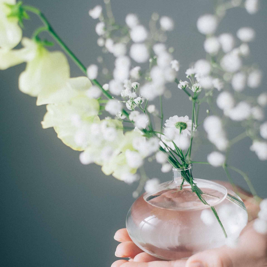 White angel's breath flowers, daisy's, and long-stemmed white flowers inside an oval shaped glass diffuser sitting inside a person's cupped hands..