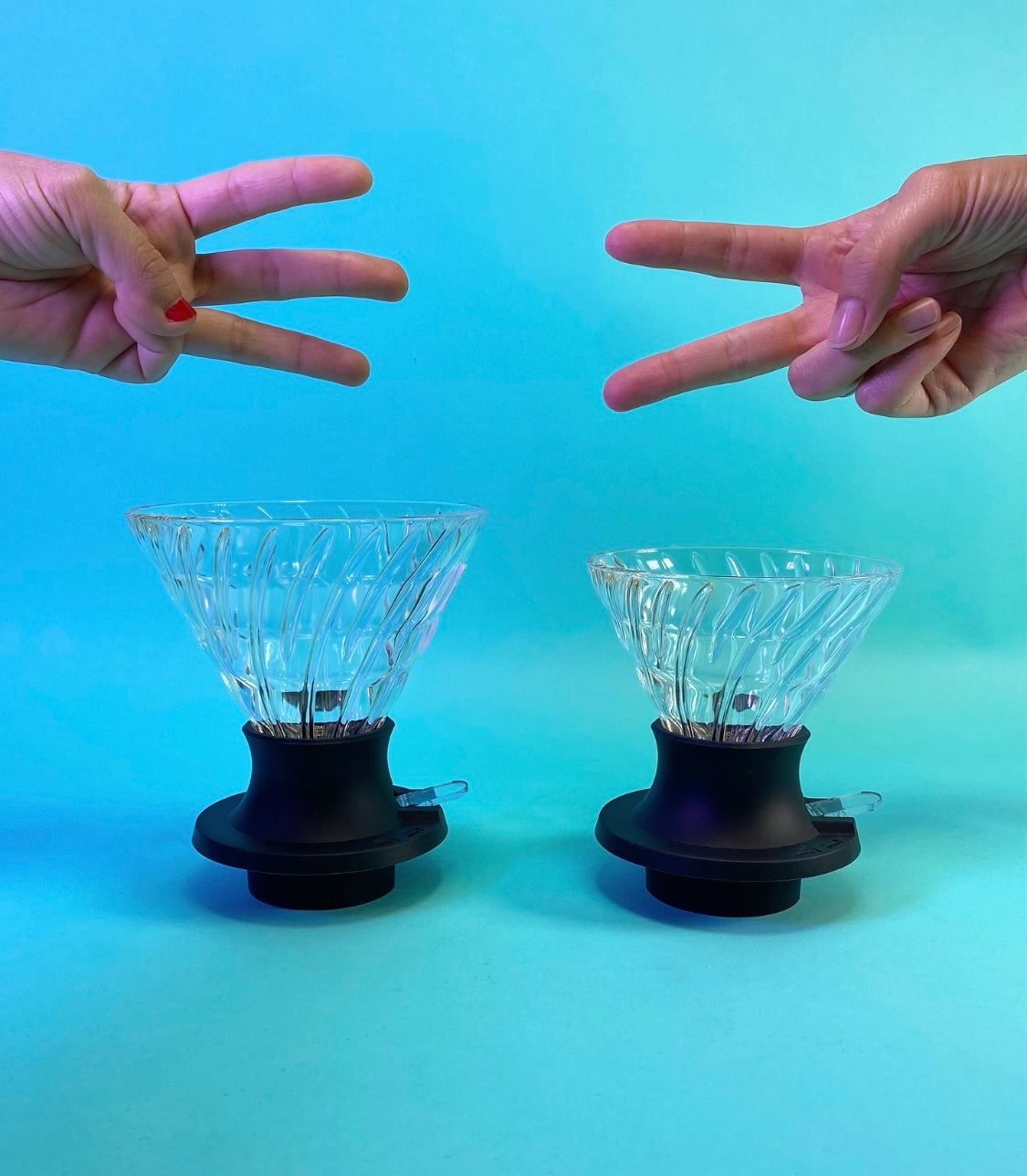 Large cone-shaped glass dripper with black silicone base with clear plastic switch, and a hand holding three fingers above to indicate size,  next to a medium cone-shaped glass dripper with black silicone base with clear plastic switch, and a hand holding two fingers above to indicate size, on a light blue background.